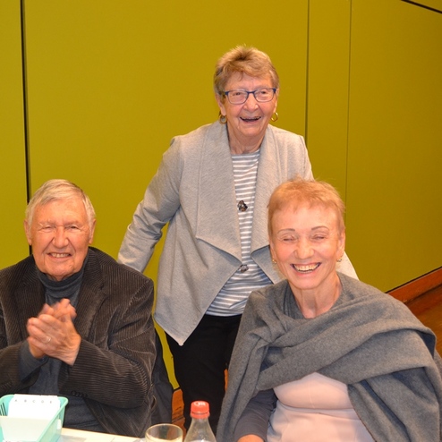 Drei &auml;ltere Menschen sitzen an einem Tisch, lachen und unterhalten sich vor einer gr&uuml;nen Wand.