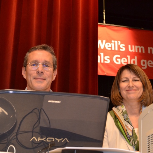 Zwei Personen l&auml;cheln hinter einem Computermonitor, mit einem roten Banner im Hintergrund.