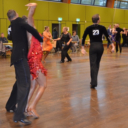 Tanzpaare in eleganter Abendkleidung zeigen einen Tanzwettbewerb auf einer Holzfl&auml;che mit Zuschauern im Hintergrund.