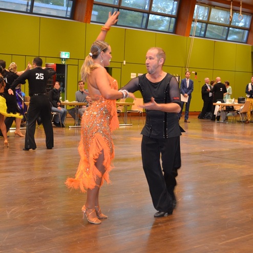 Tanzpaar in festlicher Kleidung f&uuml;hrt einen gesellschaftlichen Tanz in einem gro&szlig;en Saal auf. Zuschauer im Hintergrund.