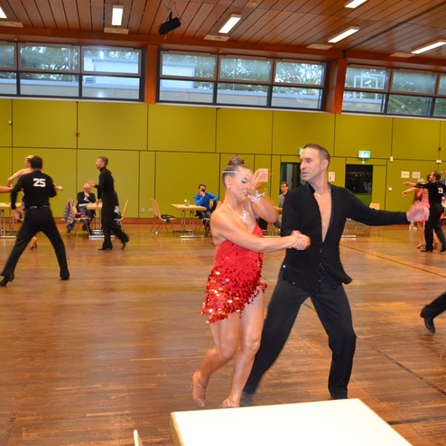 Tanzpaare in farbenfrohen Outfits tanzen auf einem Holzboden in einer Turnhalle w&auml;hrend eines Wettbewerbs.