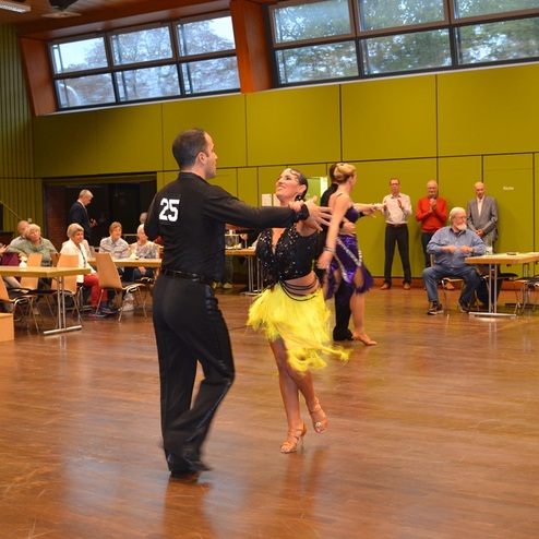 Tanzpaar in festlicher Kleidung tanzt bei einem Wettbewerb, Zuschauer sitzen an Tischen im Hintergrund.