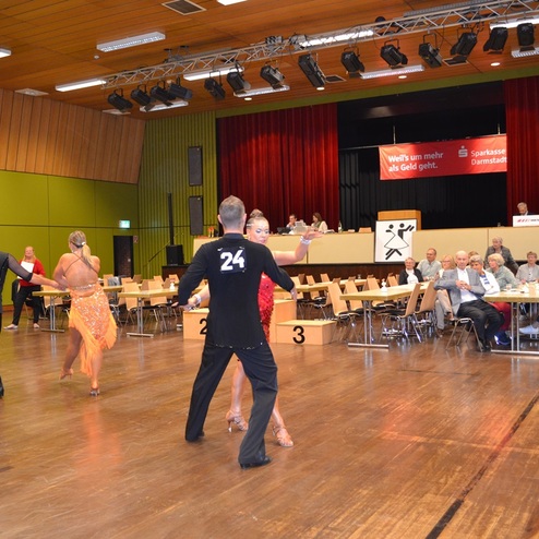 Tanzpaare in bunten Kleidern beim Auftritt in einer Turnhalle, Zuschauer beobachten an Tischen im Hintergrund.