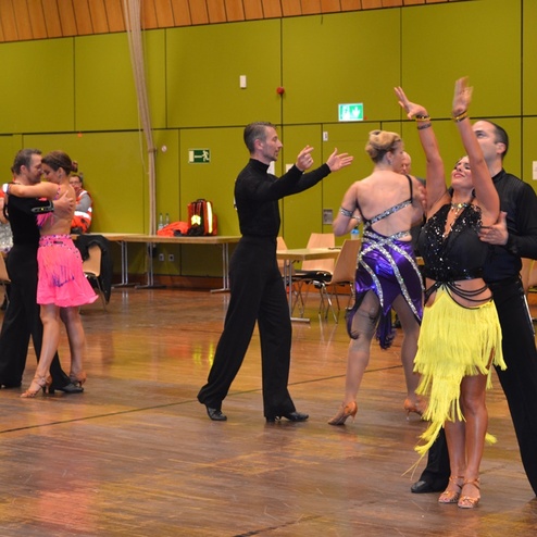 T&auml;nzerpaare in festlicher Kleidung auf einer Tanzfl&auml;che bei einer Tanzveranstaltung. St&uuml;hle und Zuschauer im Hintergrund sichtbar.