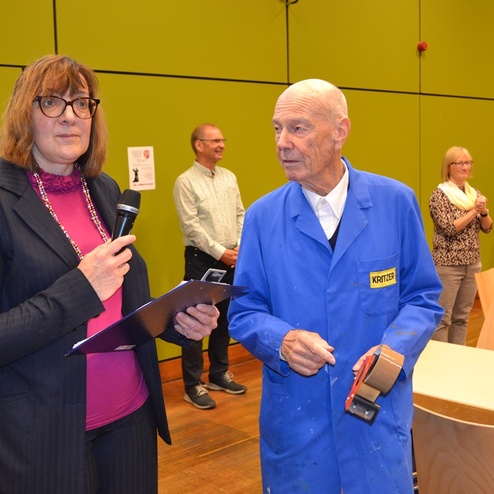 Frau mit Mikrofon spricht zu einem &auml;lteren Mann in blauer Arbeitskleidung, im Hintergrund stehen weitere Personen.