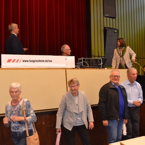 Gruppenszene in einem Veranstaltungsraum mit Menschen, die sich um eine B&uuml;hne mit Technik versammeln.