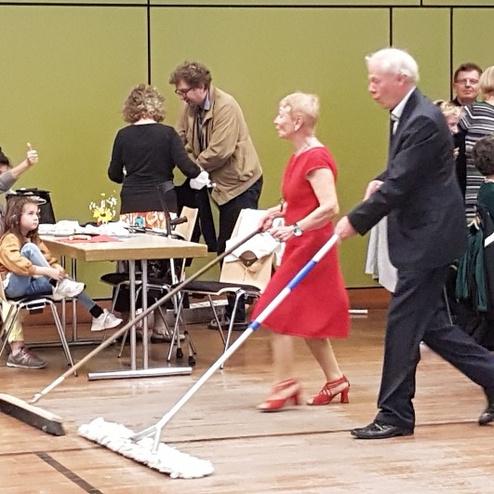 Paar in formeller Kleidung wischt mit gro&szlig;em Wischmopp &uuml;ber einen Boden in einem festlich dekorierten Raum.