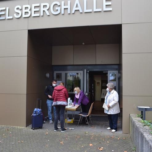 Menschen stehen vor dem Eingang zur Hegelsberghalle, einer Frau wird am Tisch etwas &uuml;berreicht.