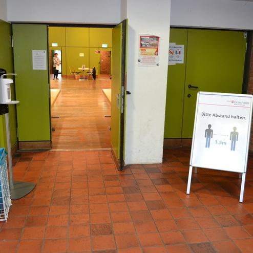 Eingang zu einem Raum mit Desinfektionsstation und Abstandshinweis auf einem Standbanner. Gr&uuml;ne W&auml;nde und Holzfu&szlig;boden sichtbar.