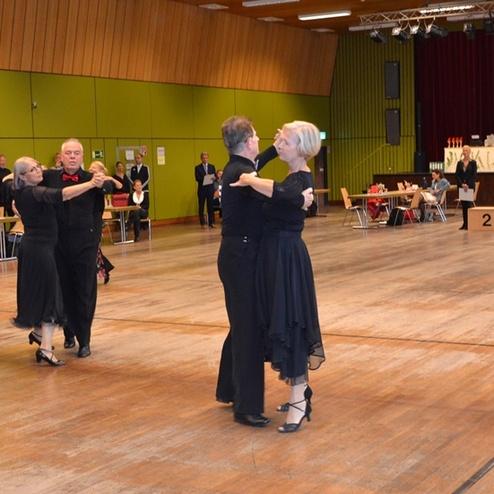 Tanzpaar in eleganter Kleidung beim Tanzen auf einer Turnierfl&auml;che; im Hintergrund Zuschauer und die Jury.
