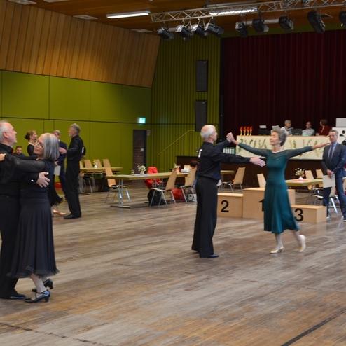 Tanzende Paare in festlicher Kleidung auf einem Parkettboden in einer farbenfrohen Veranstaltungsr&auml;umlichkeit.