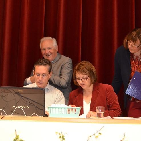 Vier Personen sitzen an einem Tisch mit einem Laptop, Unterlagen und Getr&auml;nken, vor einem rotbraunen Vorhang.