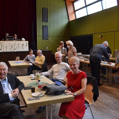 Geselliges Treffen &auml;lterer Menschen in einem Saal; Tische mit Getr&auml;nken, Blumen und Gespr&auml;chsrunden im Hintergrund.