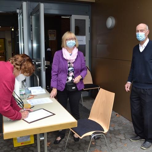Drei Personen stehen vor einem Eingang, w&auml;hrend eine Person an einem Tisch Unterlagen ausf&uuml;llt. Alle tragen Masken.