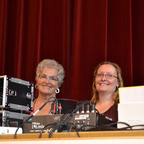 Zwei Frauen stehen l&auml;chelnd hinter einem Mischpult mit Kabeln, vor rotem Vorhang in einem Veranstaltungsraum.