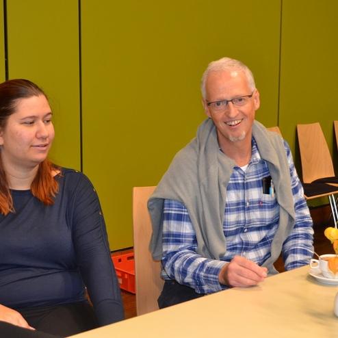 Zwei Personen sitzen an einem Tisch, eine sinniert, der andere l&auml;chelt, mit Geb&auml;ck und Blumenvasen im Vordergrund.