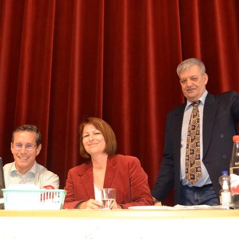 Drei Personen an einem Tisch neben einem Laptop, Getr&auml;nke und einer Pr&auml;sentationsfl&auml;che in einem Veranstaltungsraum.