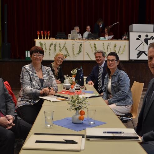 Sechsergruppe an einem Tisch mit Blumenarrangements und Papieren in einem Saal, im Hintergrund Auszeichnungen und eine B&uuml;hne.