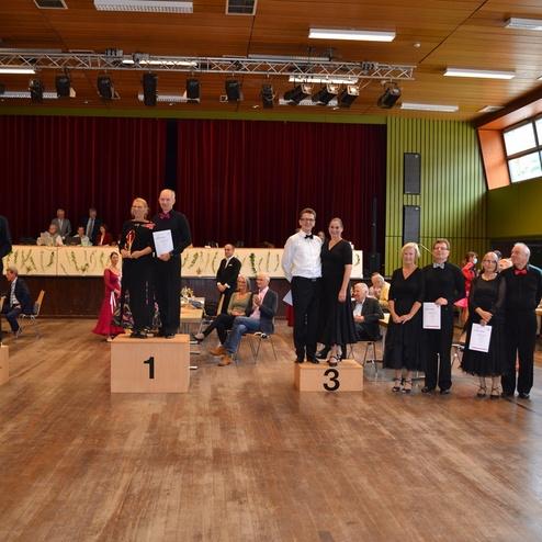 Paarweise Aufstellung der Tanzpaare auf Treppchen einer Wettbewerbsveranstaltung mit Zuschauern im Hintergrund.