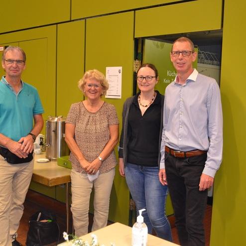 Vier Personen stehen l&auml;chelnd vor einer gr&uuml;nen Wand, im Hintergrund ist ein Tisch mit Getr&auml;nken und Snacks sichtbar.