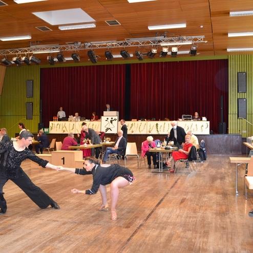 Tanzpaar in eleganter Kleidung f&uuml;hrt einen Tanz im Gro&szlig;raum mit Zuschauern und Tischen im Hintergrund auf.