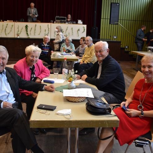 F&uuml;nf &auml;ltere Personen sitzen an einem Tisch in einem festlichen Saal, umgeben von weiteren G&auml;sten und Dekoration.