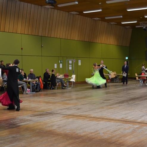Tanzpaare in eleganter Abendkleidung tanzen in einem Wettbewerbsraum mit Zuschauern an Tischen im Hintergrund.