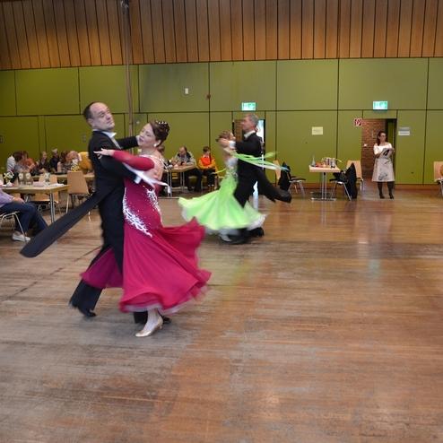 Tanzpaare in eleganter Kleidung bewegen sich &uuml;ber das Parkett eines Veranstaltungssaals, Zuschauer im Hintergrund.