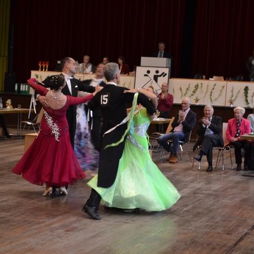 Tanzpaare in eleganten Kost&uuml;men zeigen einen Gesellschaftstanz vor Publikum in einem festlichen Saal.