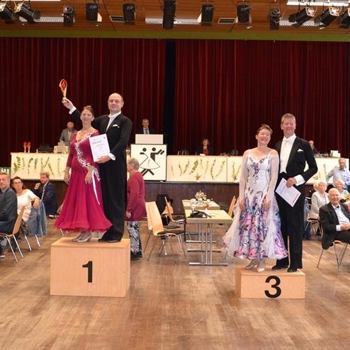 Tanzturnier mit preisgekr&ouml;nten Paaren auf dem Siegerpodest, Zuschauer im Hintergrund, festliche Atmosph&auml;re.