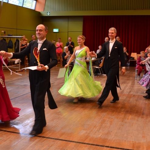 F&uuml;nf Paare in eleganter Tanzkleidung treten gemeinsam bei einem Wettbewerb auf einer Holzfl&auml;che auf.
