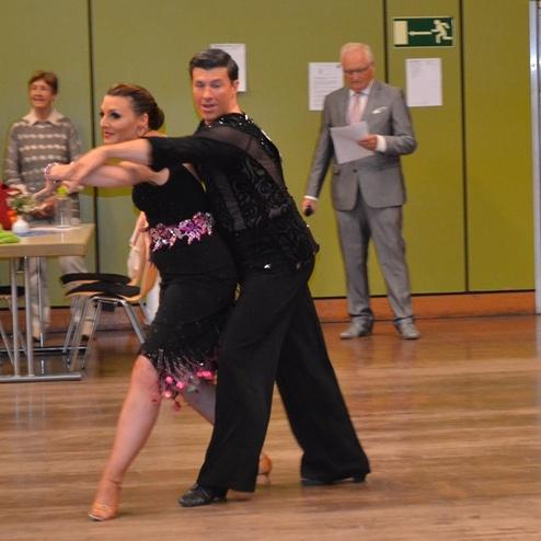 Tanzende Paare in eleganter Kleidung auf einem Parkettboden, w&auml;hrend Zuschauer im Hintergrund zuschauen.