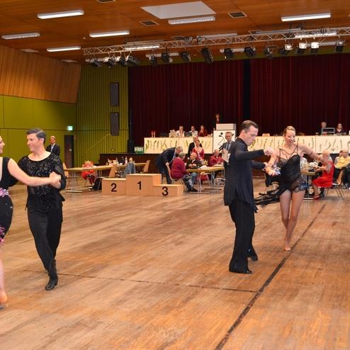 Tanzpaare in eleganter Kleidung tanzen in einem gro&szlig;en Saal vor einer Zuschauergruppe und einer Jury.