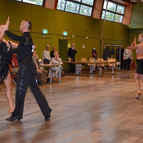 Tanzende Paare in eleganter Kleidung w&auml;hrend eines Wettbewerbs, umgeben von Zuschauern in einem festlichen Saal.