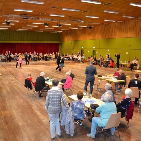 Tanzveranstaltung in einem gro&szlig;en Saal mit zahlreichen G&auml;sten, die an Tischen sitzen, w&auml;hrend Paare tanzen.