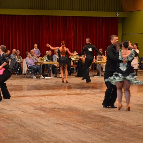 Tanzpaare in eleganter Kleidung f&uuml;hren einen T&auml;nz in einem Saal vor einemPublikum durch.