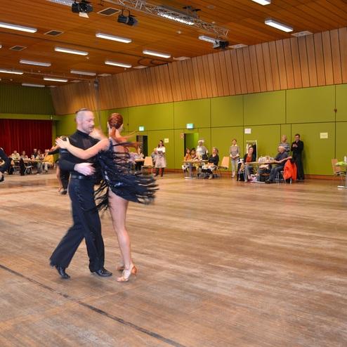 Tanzpaare in festlicher Kleidung tanzen auf einer gl&auml;nzenden Tanzfl&auml;che, Zuschauer sitzen an Tischen im Hintergrund.