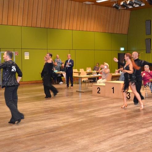 Tanzpaare in festlicher Kleidung treten auf einer B&uuml;hne w&auml;hrend eines Wettbewerbs auf, Zuschauer sitzen an Tischen.