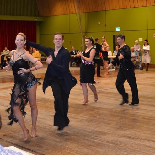 Tanzende Paare in festlicher Kleidung auf einem Parkettboden w&auml;hrend einer Tanzveranstaltung. Zuschauer beobachten im Hintergrund.
