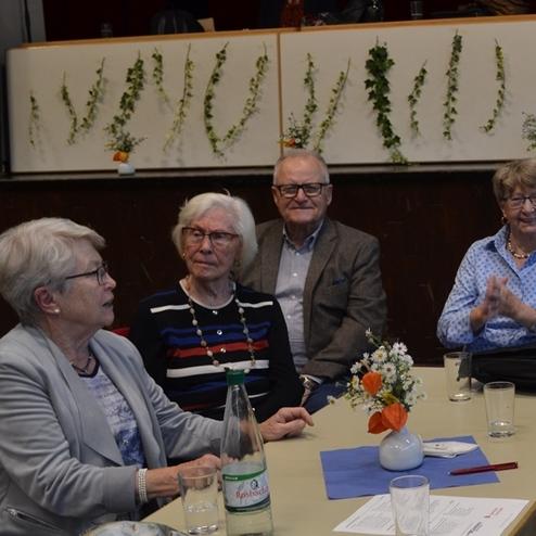 Vier &auml;ltere Personen sitzen um einen Tisch, dekoriert mit Blumen und Getr&auml;nken, in einem festlichen Raum.