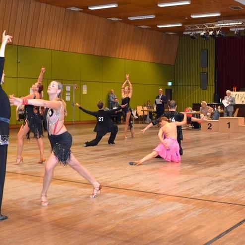 Tanzwettbewerb mit Paaren in schicken Kost&uuml;men auf einer Holztanzfl&auml;che, Zuschauer sitzen an Tischen im Hintergrund.