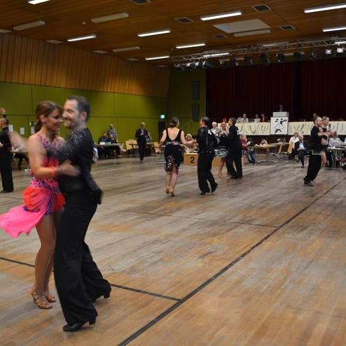 Paare tanzen in einem Wettbewerb auf einer Holzb&uuml;hne, Zuschauer sitzen an Tischen im Hintergrund.