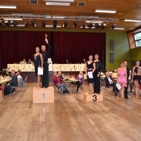 T&auml;nzerpaar auf Siegertreppe bei einem Wettbewerb, zahlreiche Zuschauer sitzen an Tischen im Hintergrund.