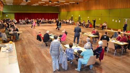 Tanzveranstaltung in einem großen Saal mit zahlreichen Gästen, die an Tischen sitzen, während Paare tanzen.