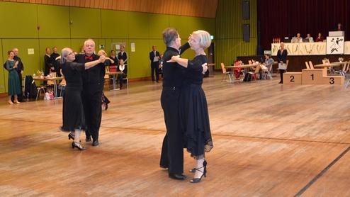 Tanzpaar in eleganter Kleidung beim Tanzen auf einer Turnierfläche; im Hintergrund Zuschauer und die Jury.