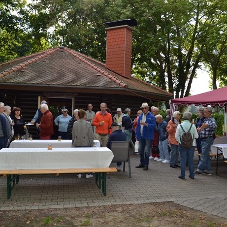 Gruppe von Menschen versammelt sich vor einem Holzhaus und einem roten Zelt, umgeben von Bäumen und Tischen.