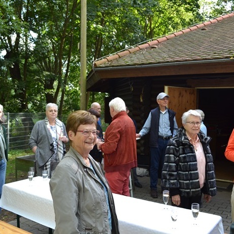 Gruppe von Menschen steht draußen vor einer Holzlaube, einige halten Glas Getränke in der Hand, Bäume im Hintergrund.