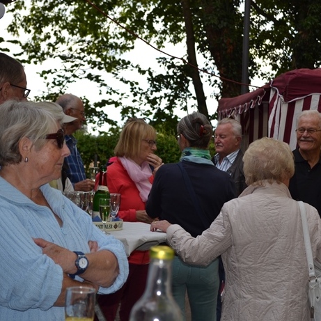 Gruppe älterer Menschen unterhält sich an einem Stehtisch, umgeben von Bäumen und einem festlich dekorierten Zelt.