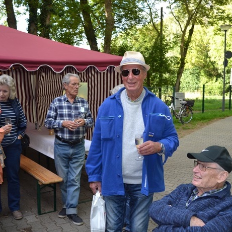Gruppe älterer Menschen beim geselligen Beisammensein in einem Park, umgeben von Bäumen und einem Stand.