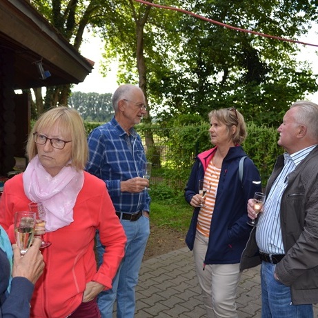 Gruppengespräch von fünf Personen im Freien, mit Getränken in der Hand, umgeben von Bäumen und einer Holzstruktur.